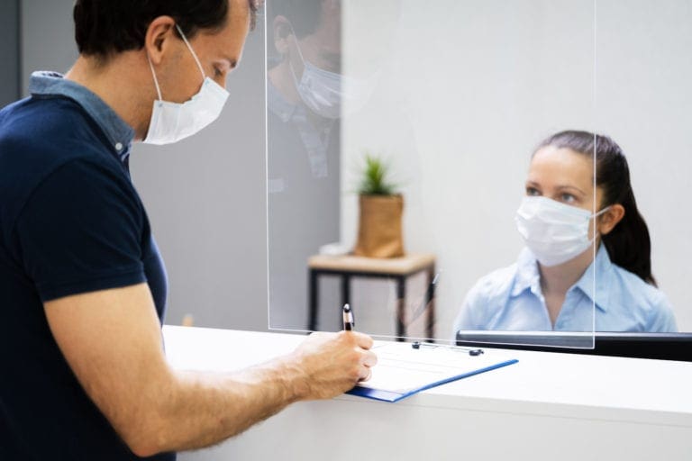 Client Signing Paper At Reception Desk In Face Mask