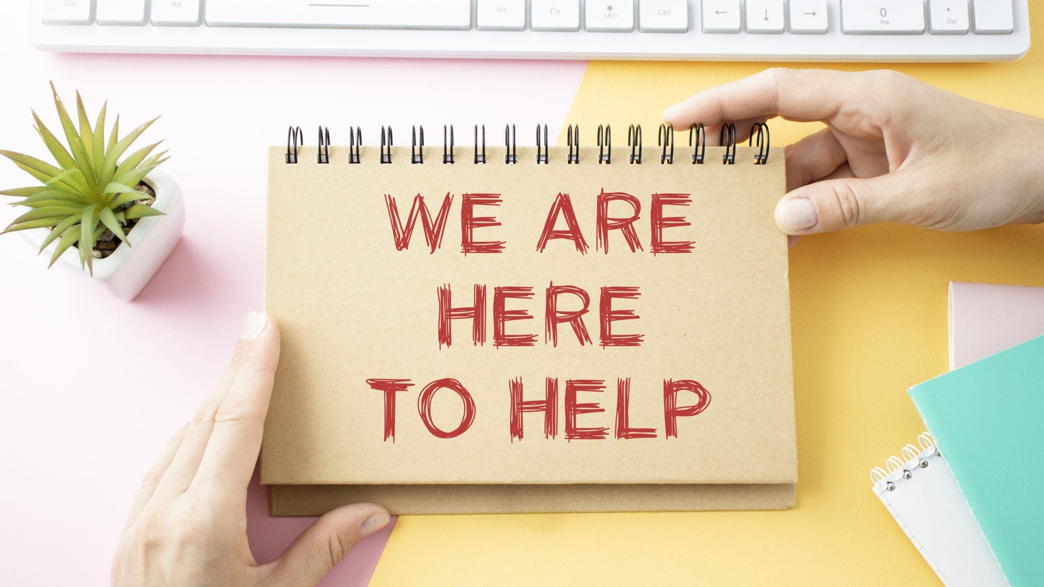 Spiral notepad with red text reading "We Are Here To Help" held by two hands, next to a plant and a keyboard on a colourful desk
