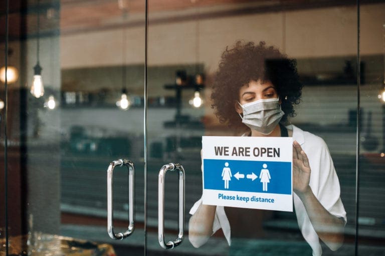 Business owner wearing a mask places a “We Are Open” social distancing sign on the shop door