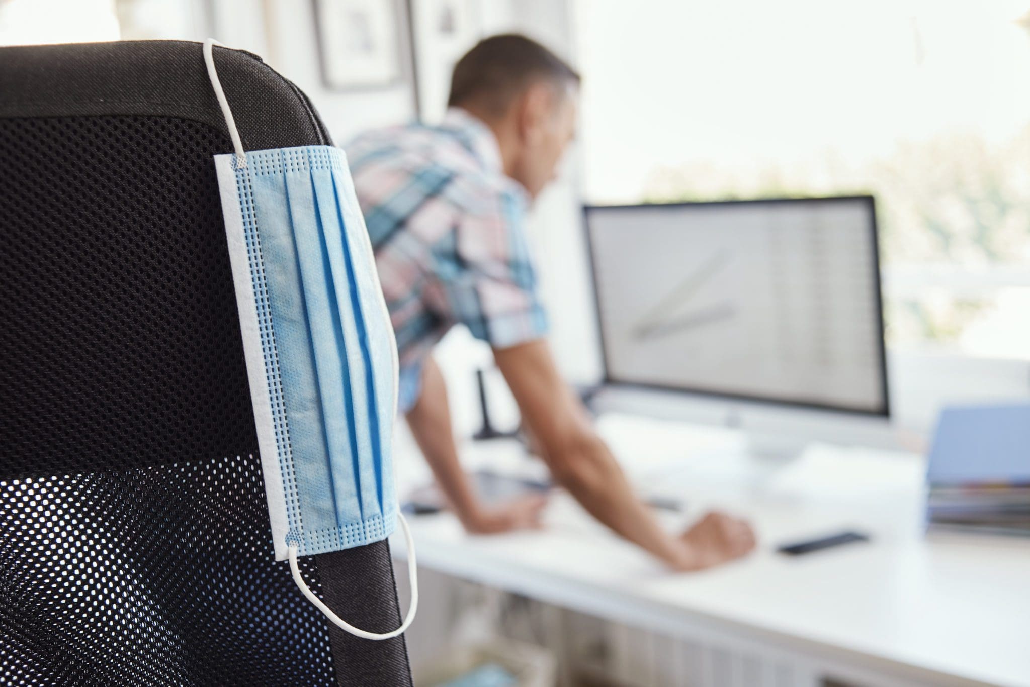 closeup of a blue face mask hanging from the back of an office chair and a young man checking something in the screen