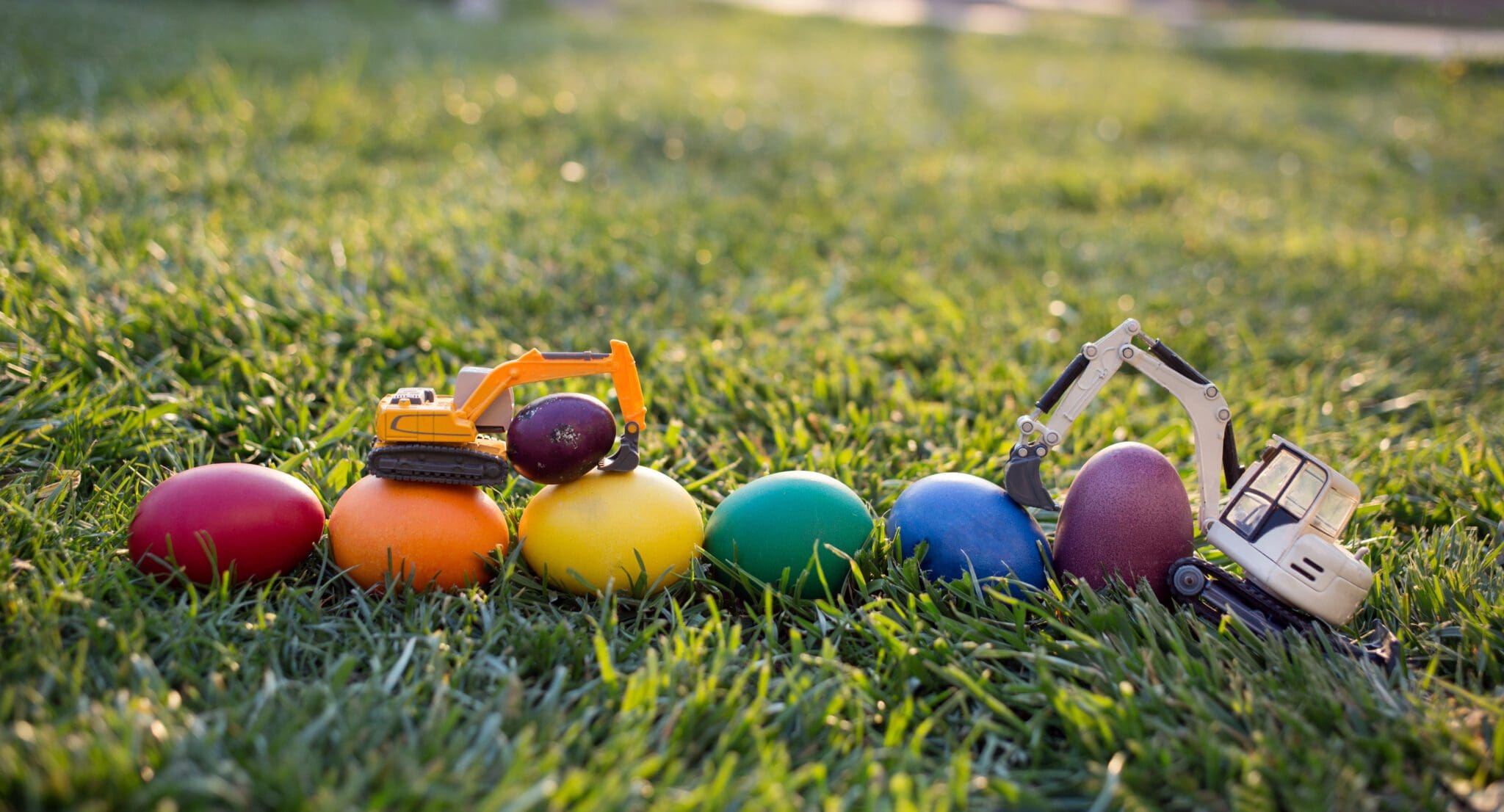 Toy diggers moving colourful Easter eggs arranged in a row on green grass, representing renewal and creativity.