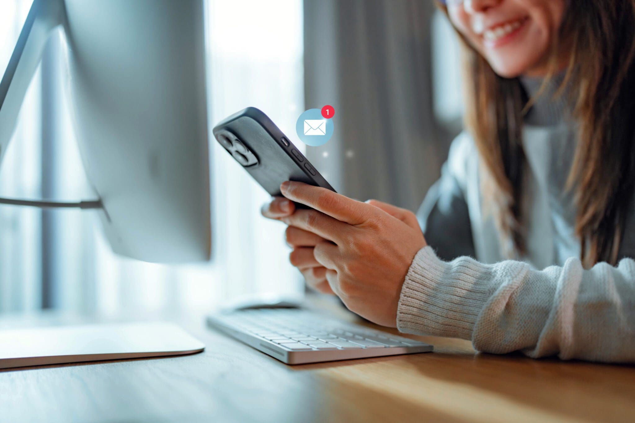 A woman checking her smartphone for a new email while sitting at a desk with a computer in a bright workspace.