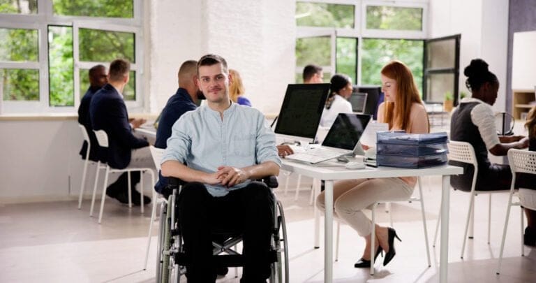 A person in a wheelchair working in a modern office with a diverse group of colleagues, reflecting O'Brien Media's commitment to being a Disability Confident Leader and promoting inclusive workplaces.