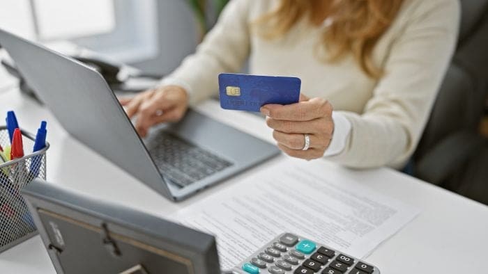 Person holding a blue bank card while entering payment details on a laptop at a desk