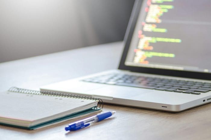 Laptop displaying source code next to a notebook and pen on a desk, representing web development planning
